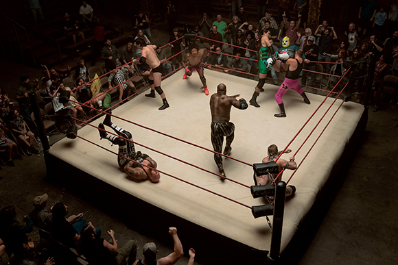 Wrestlers in a ring performing moves before a seated crowd in a dimly lit venue.