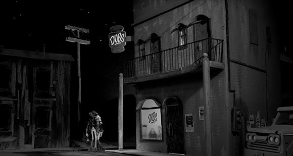 Black and white shot of a western town street at night with a neon sign reading 'DUBS' above a building.