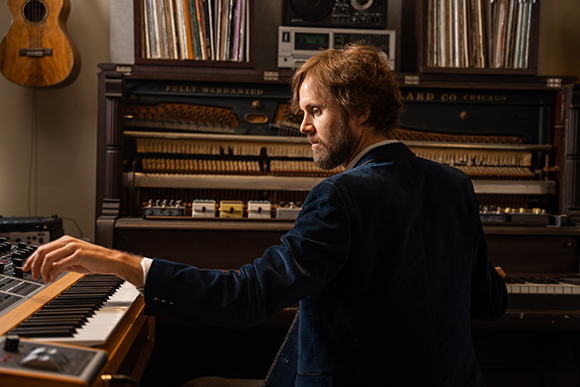 A man in a dark blazer plays a synthesizer in front of a large, vintage upright piano.