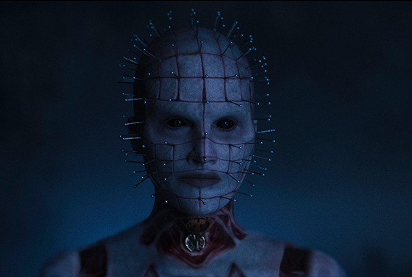 A pale humanoid figure with a grid of lines on their face and metal pins sticking out of their head and skin.