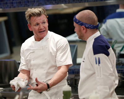 A chef in a white jacket gestures while speaking to another chef wearing a blue headband in a kitchen.