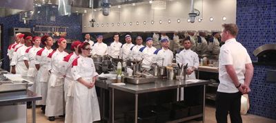 A group of chefs in white uniforms stand in a professional kitchen listening to a man in a white chef's coat.