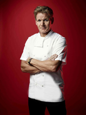 A man with short blond hair wearing a white chef's coat and black pants, posing with arms crossed against a red backdrop.