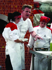 A male chef in white uniform shouting while pointing at a pan held by a female cook in a kitchen.