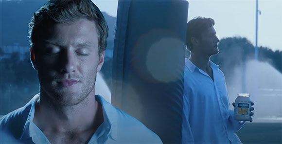 A man with closed eyes in the foreground and a man in a white shirt holding a supplement jar in the background.