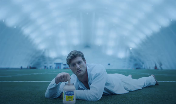 A man in white clothing lies on a green field, holding a jar of Hellmann's mayonnaise in a large, blue-lit dome.