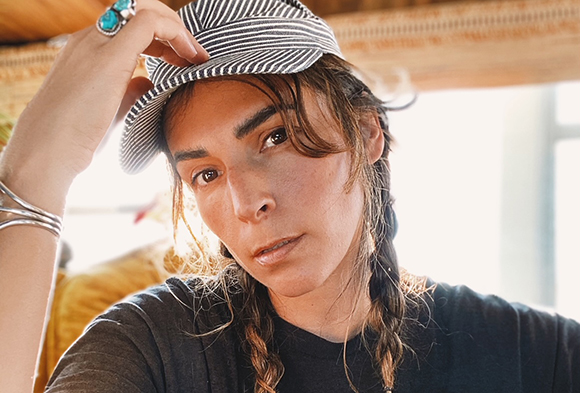A person with braided hair wearing a striped cap and a dark t-shirt looks toward the camera.