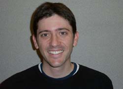 A smiling man with short dark hair wearing a black crewneck shirt against a plain gray background.