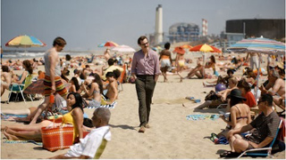 A man in a purple shirt and dark trousers walks through a crowded, sunny beach filled with people and umbrellas.