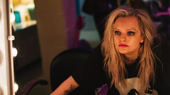 A blonde woman in a black t-shirt looks into a lit vanity mirror with a serious expression.