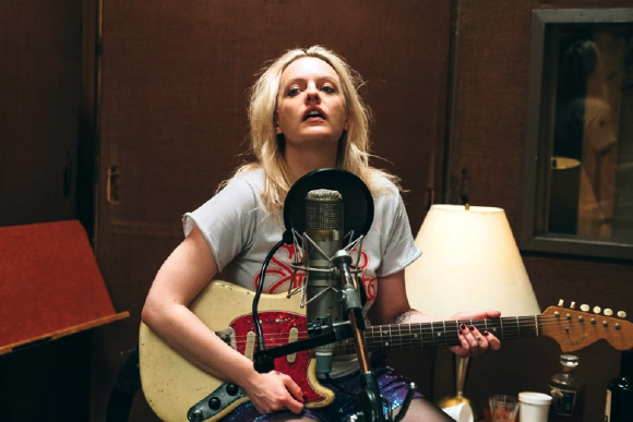 A blonde woman in a grey t-shirt sings into a studio microphone while playing a cream electric guitar.