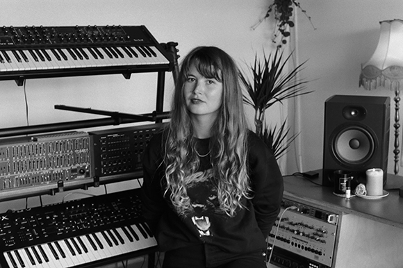 A woman sits in a music studio surrounded by electronic keyboards, synthesizers, and a studio monitor.