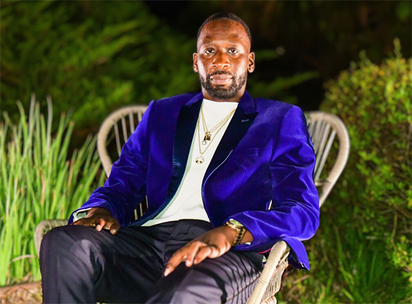 A man wearing a blue velvet blazer and white shirt sits in a wicker chair outdoors at night.