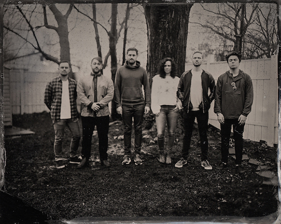 Six people stand in a row in a yard against a backdrop of trees and a wooden fence.