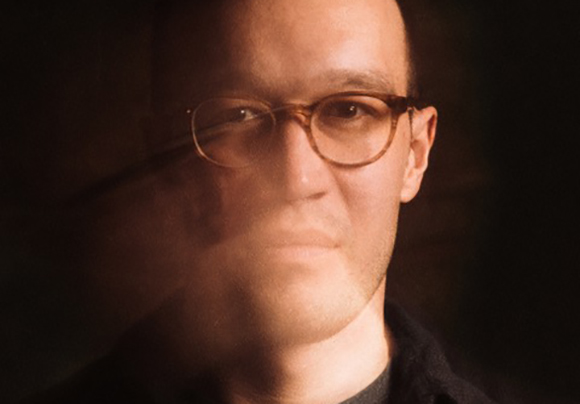 A man with glasses looks forward in a dimly lit, warm-toned portrait.