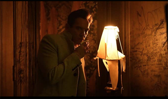 A man in a light-colored suit stands in a dark room next to a glowing lamp.