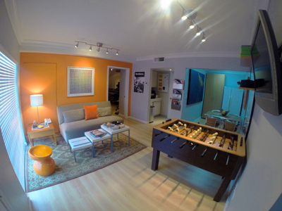 A living room with an orange wall, a grey sofa, and a foosball table on a light wood floor.