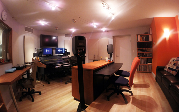 A music recording studio with a mixing console, speakers, keyboards, and a wooden desk.