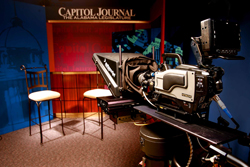 A professional video camera on a tripod in a studio with two empty chairs and a Capitol Journal sign.