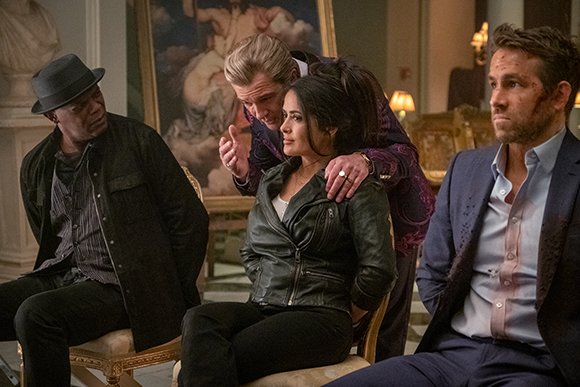 Four people sit in a room, including a man in a blue suit and a woman in a black leather jacket.