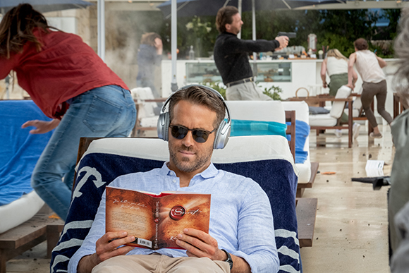 A man in sunglasses relaxes with headphones and a book while people run and a man points a gun in the background.