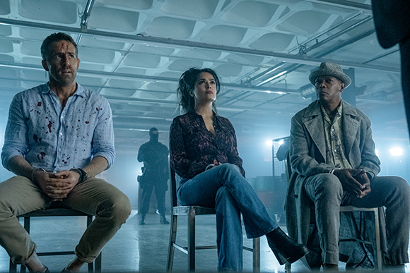 Three people sit in chairs in a blue-toned, industrial room with a figure in the background.