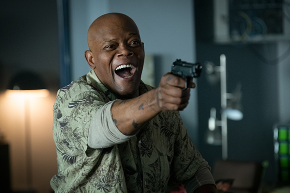 A bald man in a patterned shirt laughs while pointing a handgun toward the camera.