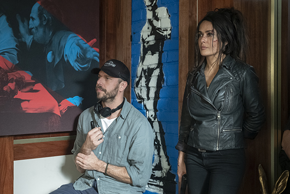 A man in a grey shirt and cap sits next to a woman in a black leather jacket in front of large paintings.