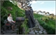 A person in a dark wizard costume stands in a lush green garden near a person sitting on a bench.