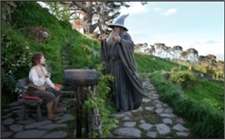 A wizard in grey robes points a staff at a man sitting on a bench in a lush, hilly garden.