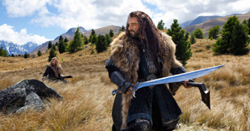 A man in dark clothing and fur trim kneels in a field holding a large sword against a mountain backdrop.