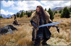 A man in fur-lined armor kneels in a field holding a large sword, with mountains and trees in the background.
