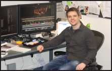A man in a dark shirt and jeans sits at a desk with two large computer monitors in an office setting.