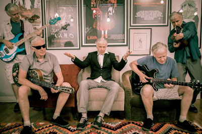 Four men sit and stand with guitars and a bass in a room decorated with framed pictures.