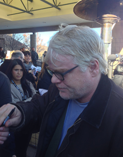 Philip Seymour Hoffman with blond hair and glasses signs an autograph for someone off camera.