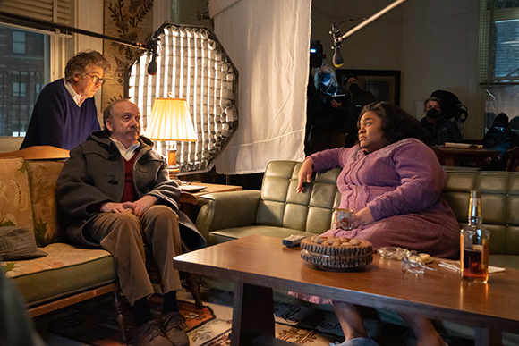 Actors sit on a couch while a camera operator and lighting equipment are visible in a studio setting.