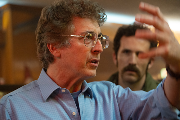 A man wearing glasses gestures with an outstretched hand while looking to the side.