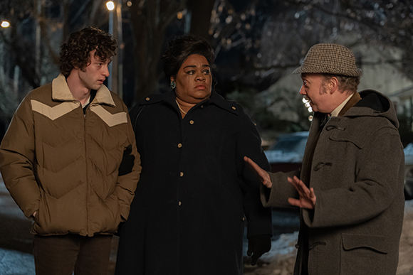 A man in a brown jacket, a woman in a black coat, and a man in a grey coat talking outdoors at night.