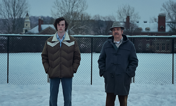 Two men stand in a snowy field behind a chain-link fence with buildings in the background.