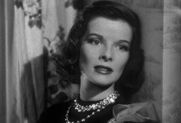Black and white close-up of a woman with wavy hair wearing a dark top and sparkling necklaces.