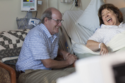 A man in a blue checkered shirt sits next to a woman laughing in a hospital bed.