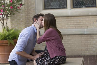 A man in a light blue shirt and a woman in a pink sweater kiss while sitting outdoors.