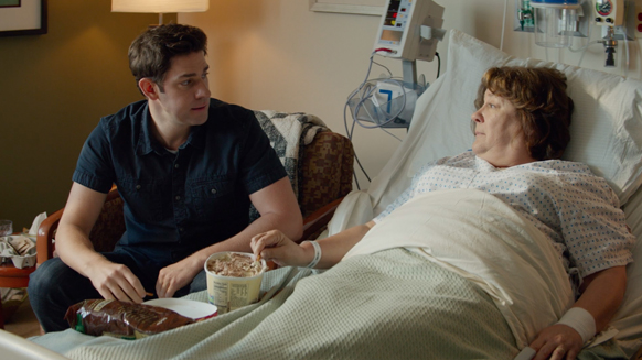 A young man in a dark shirt sits beside a woman lying in a hospital bed.