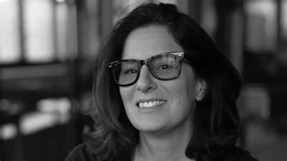 A smiling woman with dark hair and glasses looks toward the camera in a black and white portrait.