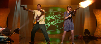 Two actors on a stage holding prop guns in front of a green star-shaped backdrop.