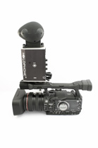 A black professional video camera with a large microphone and spherical windjammer mounted on top.