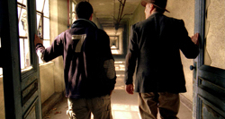Two men seen from behind standing in a doorway, looking down a long, sunlit corridor.