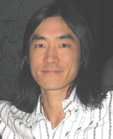A man with long dark hair and a slight smile wearing a white patterned shirt.