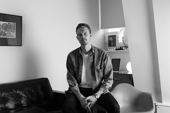 A man in a button-down shirt and t-shirt sits facing the camera in a room.