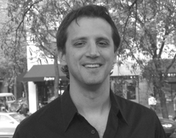 A smiling man with dark hair wearing a dark button-down shirt in an outdoor setting.